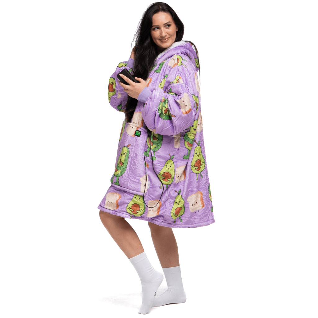 Eine Person trägt einen lilafarbenen, übergroßen Kapuzenpullover von The Hoti, der mit Avocado-Illustrationen verziert ist, steht lächelnd da und hält ein Smartphone in der Hand. Sie hat langes dunkles Haar und trägt weiße Socken.