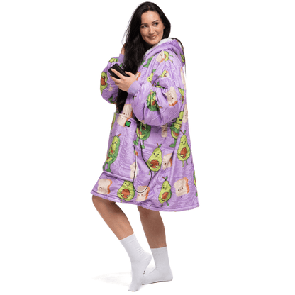 Eine Person trägt einen lilafarbenen, übergroßen Kapuzenpullover von The Hoti, der mit Avocado-Illustrationen verziert ist, steht lächelnd da und hält ein Smartphone in der Hand. Sie hat langes dunkles Haar und trägt weiße Socken.