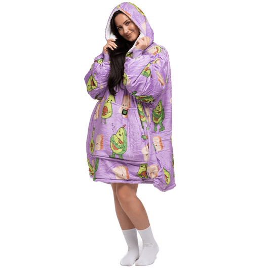 Eine Person trägt einen übergroßen Avokado Familie Hoti-Kapuzenpullover in Lila mit Avocado- und Toastmustern der Marke The Hoti. Sie hat lange Haare und posiert mit einem verspielten Lächeln, die Hände vor dem Gesicht. Vor dem schlichten weißen Hintergrund sind weiße Socken zu sehen.