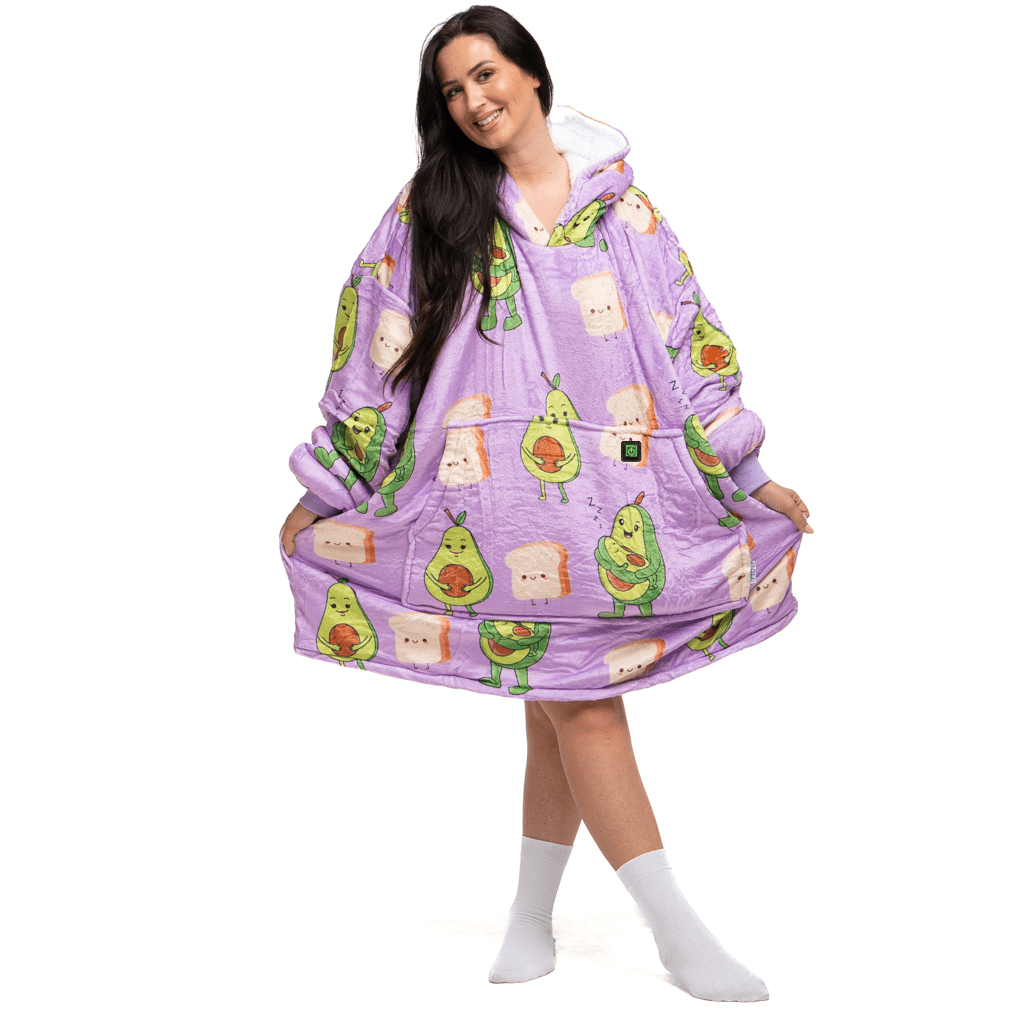 Eine Frau trägt einen großen, gemütlichen lila Kapuzenpullover aus der Kollektion Avokado Familie Hoti von The Hoti mit Avocado-Cartoons und Toast. Sie steht lächelnd da, zieht mit den Händen die Ränder des Kapuzenpullovers nach außen und trägt weiße Socken.