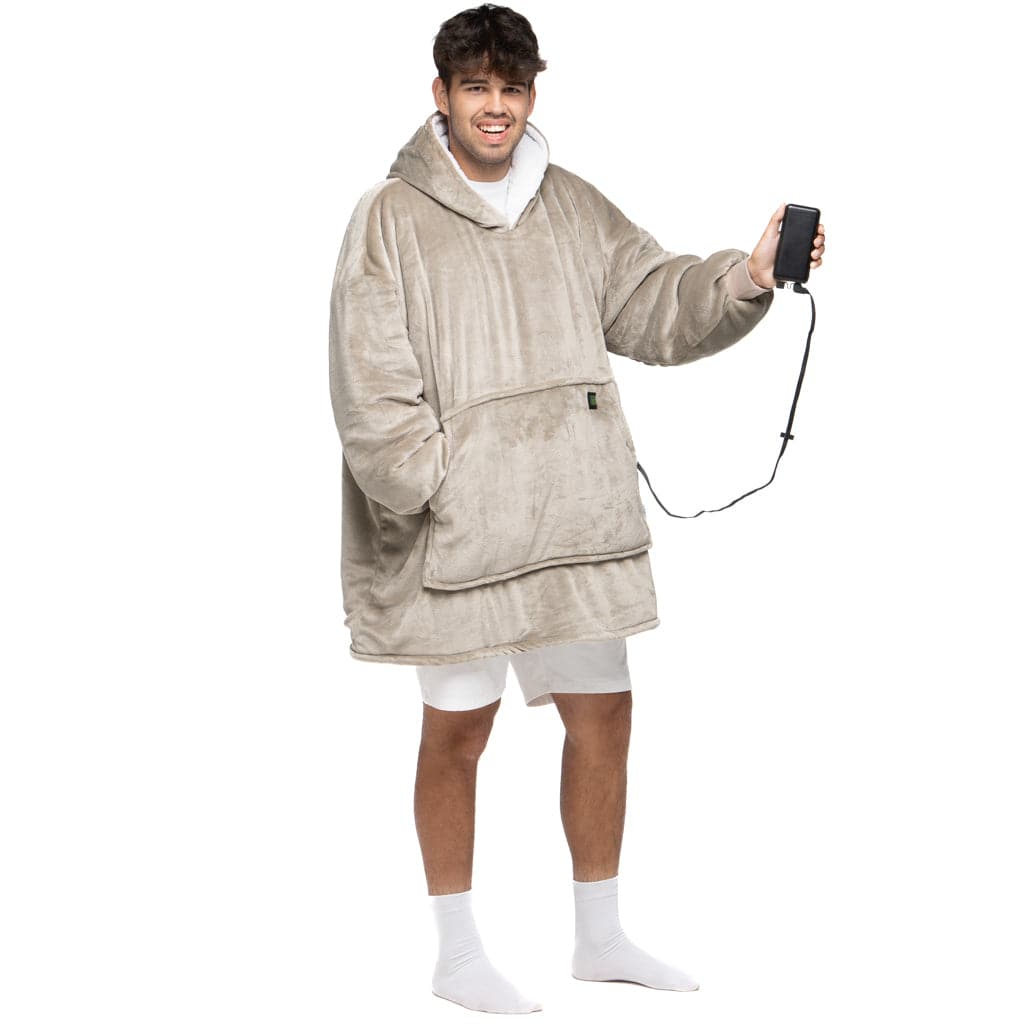 Eine lächelnde Person trägt den Flauschiger Beige Hoti von The Hoti, einen übergroßen Fleece-Hoodie mit einer großen Vordertasche, dazu weiße Shorts und Socken. Sie hält ein schwarzes Telefon, das an ein Kabel angeschlossen ist, und steht vor einem schlichten weißen Hintergrund.