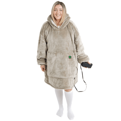 Eine Person lächelt, während sie den Flauschiger Beige Hoti von The Hoti trägt, eine übergroße Kapuzendecke aus weichem grauem Material mit einer großen Vordertasche. Sie hält ein kabelgebundenes Gerät in der Hand und trägt lange weiße Socken.