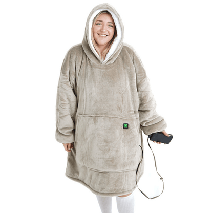 Eine Person trägt einen großen, gemütlichen Flauschiger Beige Hoti von The Hoti mit flauschiger Textur und Vordertasche und lächelt, während sie ein schwarzes Smartphone hält. Die Kapuzendecke bedeckt die Person vollständig und ähnelt einem übergroßen Pullover.