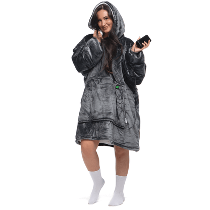 Eine Person, eingehüllt in einen bequemen Kapuzenmantel „Grauer Zauber Hoti“ von The Hoti, hält ein Smartphone. Sie steht vor einem weißen Hintergrund, lächelt und trägt weiße Socken.