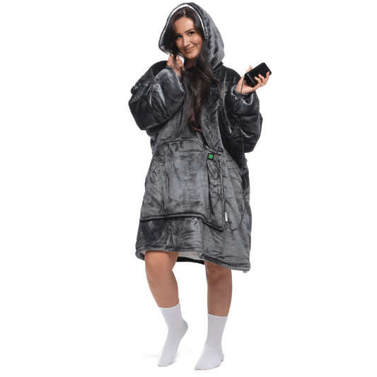 Eine Person, eingehüllt in einen bequemen Kapuzenmantel „Grauer Zauber Hoti“ von The Hoti, hält ein Smartphone. Sie steht vor einem weißen Hintergrund, lächelt und trägt weiße Socken.