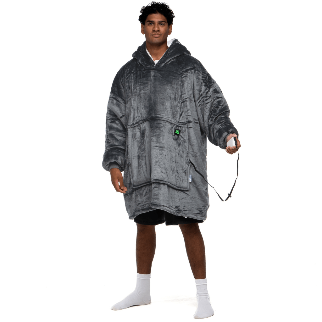 Eine Person, die den Grauer Zauber Hoti trägt, eine große, flauschige Kapuzendecke von The Hoti, hält einen Gamecontroller. Auf einem Bein stehend und mit weißen Socken bekleidet, strahlt sie eine entspannte und lässige Stimmung aus.