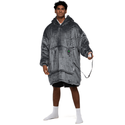 Eine Person, die den Grauer Zauber Hoti trägt, eine große, flauschige Kapuzendecke von The Hoti, hält einen Gamecontroller. Auf einem Bein stehend und mit weißen Socken bekleidet, strahlt sie eine entspannte und lässige Stimmung aus.