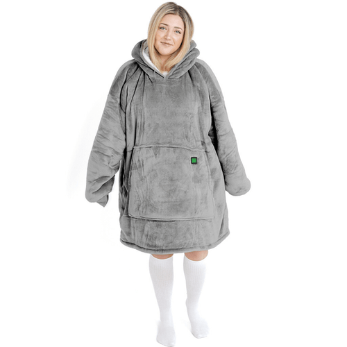 Eine Person ist in das silberne Kuschel Hoti von The Hoti gehüllt, eine große, gemütliche graue Kapuzendecke mit einer übergroßen Tasche, in der die Hände ruhen. Sie trägt außerdem weiße Socken und hat vor einem schlichten weißen Hintergrund ein sanftes Lächeln im Gesicht.