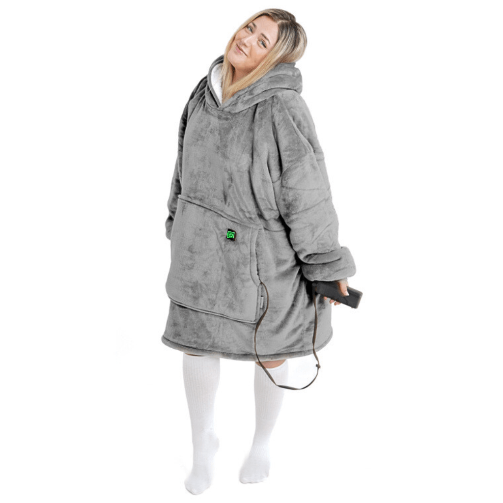 Eine Person trägt den Silber Kuschel Hoti, ein übergroßes graues Kapuzensweatshirt von The Hoti, mit einer Vordertasche und hält eine Fernbedienung in der Hand. Das plüschige und weiche Sweatshirt unterstreicht ihr entspanntes und bequemes Aussehen, während sie vor einem weißen Hintergrund steht.