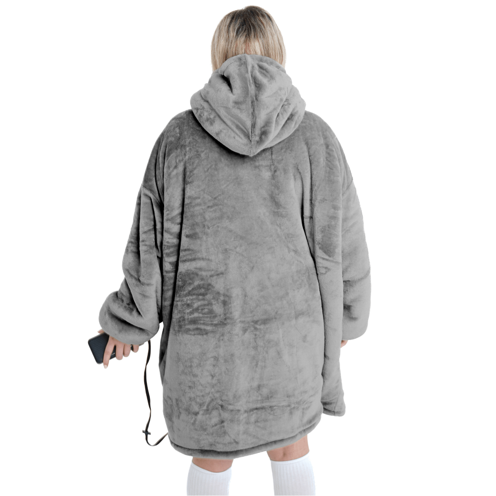 Eine Person, die die übergroße, graue Silber Kuschel Hoti von The Hoti trägt, steht mit dem Rücken zur Kamera und hält ein Smartphone in der Hand. Diese Plüschdecke sieht unglaublich gemütlich aus, ergänzt durch kniehohe weiße Socken. Der Hintergrund ist schlicht weiß.