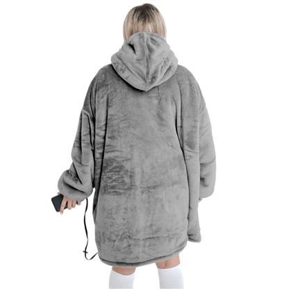 Eine Person, die die übergroße, graue Silber Kuschel Hoti von The Hoti trägt, steht mit dem Rücken zur Kamera und hält ein Smartphone in der Hand. Diese Plüschdecke sieht unglaublich gemütlich aus, ergänzt durch kniehohe weiße Socken. Der Hintergrund ist schlicht weiß.