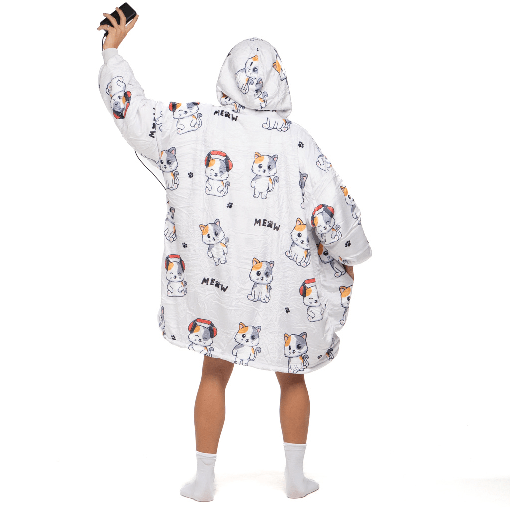 Eine Person trägt „The Hoti’s Träumende Katze Hoti“, eine gemütliche, übergroße Kapuzendecke mit Cartoon-Katzenillustrationen und dem Wort „MEOW“, und hält ein Smartphone hoch. Sie trägt weiße Socken und steht vor einem weißen Hintergrund.