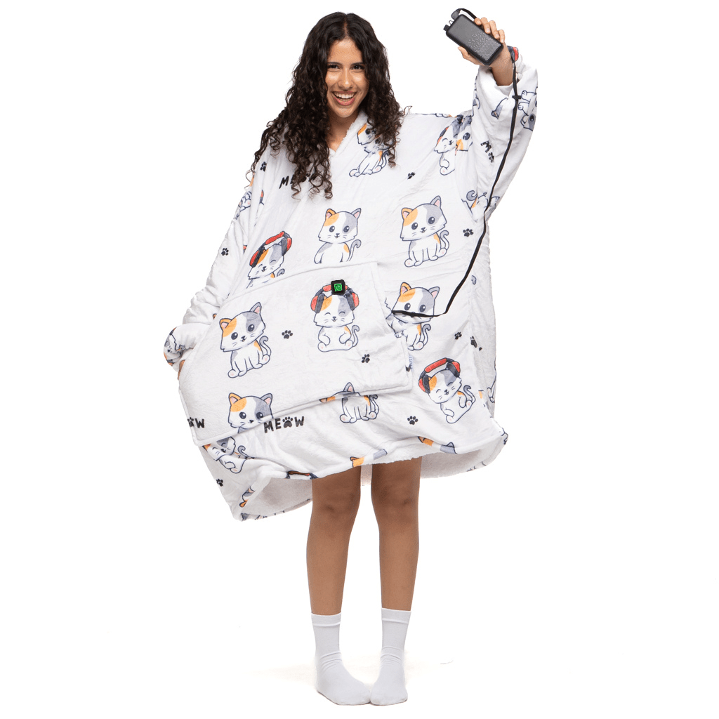 Eine Person trägt eine bequeme, übergroße tragbare Decke „Träumende Katze Hoti“ mit einem bezaubernden Muster aus Katzen mit Kopfhörern und dem Wort „Meow“ der Marke The Hoti, hält ein Smartphone und lächelt, dazu weiße Socken.