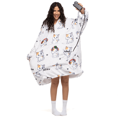 Eine Person trägt eine bequeme, übergroße tragbare Decke „Träumende Katze Hoti“ mit einem bezaubernden Muster aus Katzen mit Kopfhörern und dem Wort „Meow“ der Marke The Hoti, hält ein Smartphone und lächelt, dazu weiße Socken.