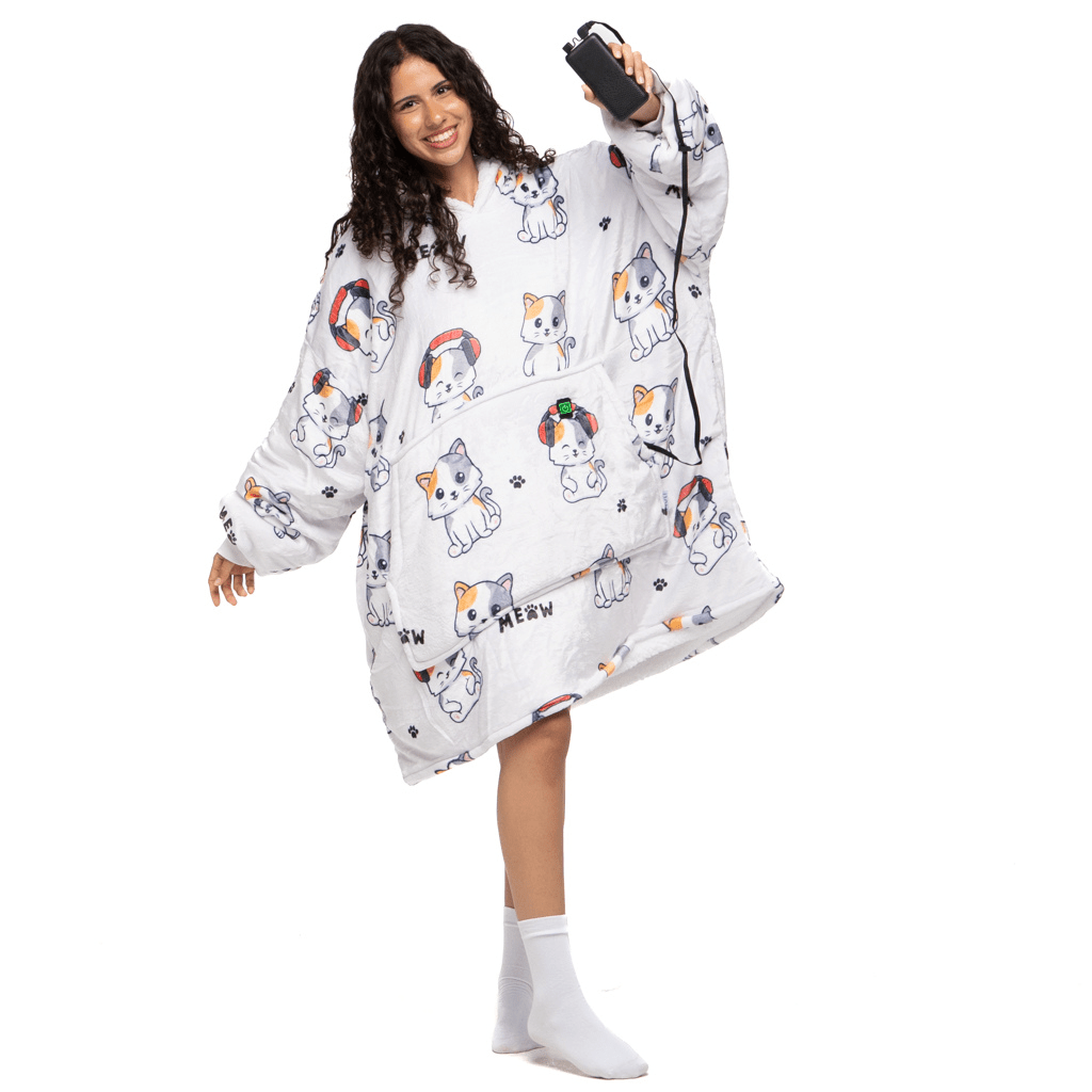 Eine Person mit langen lockigen Haaren trägt fröhlich einen übergroßen Träumende Katze Hoti-Kapuzenpullover von The Hoti. Sie lächelt, hält einen Gaming-Controller und balanciert auf einem Bein mit weißen Socken vor einem schlichten Hintergrund.