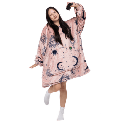 Eine Person, die den übergroßen Kapuzenpullover „Träumender Stern“ von The Hoti trägt, der mit einem himmlischen Muster in Pink verziert ist, lächelt und macht eine verspielte Pose, während sie ein Telefon hält. Sie trägt weiße Socken und steht auf einem Bein, das andere ist leicht angehoben.