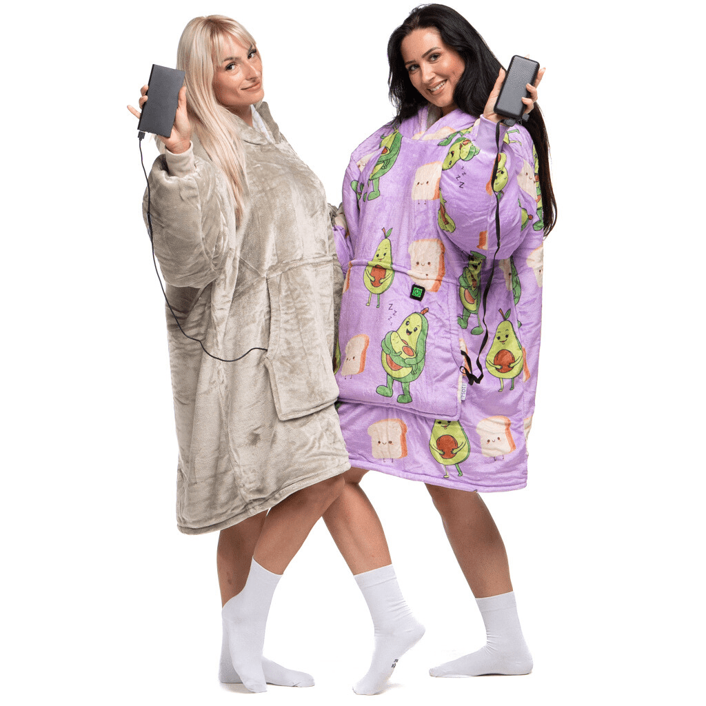 Zwei Menschen lächeln, eingehüllt in übergroße tragbare Decken von The Hoti, jede ausgestattet mit batteriebetriebenen Heizkissen, dem sogenannten Hoti Power-Paket. Eine Decke ist beige, die andere lila und mit Avocado- und Toastmustern verziert. Sie halten ihre Powerbanks an den Decken angeschlossen.